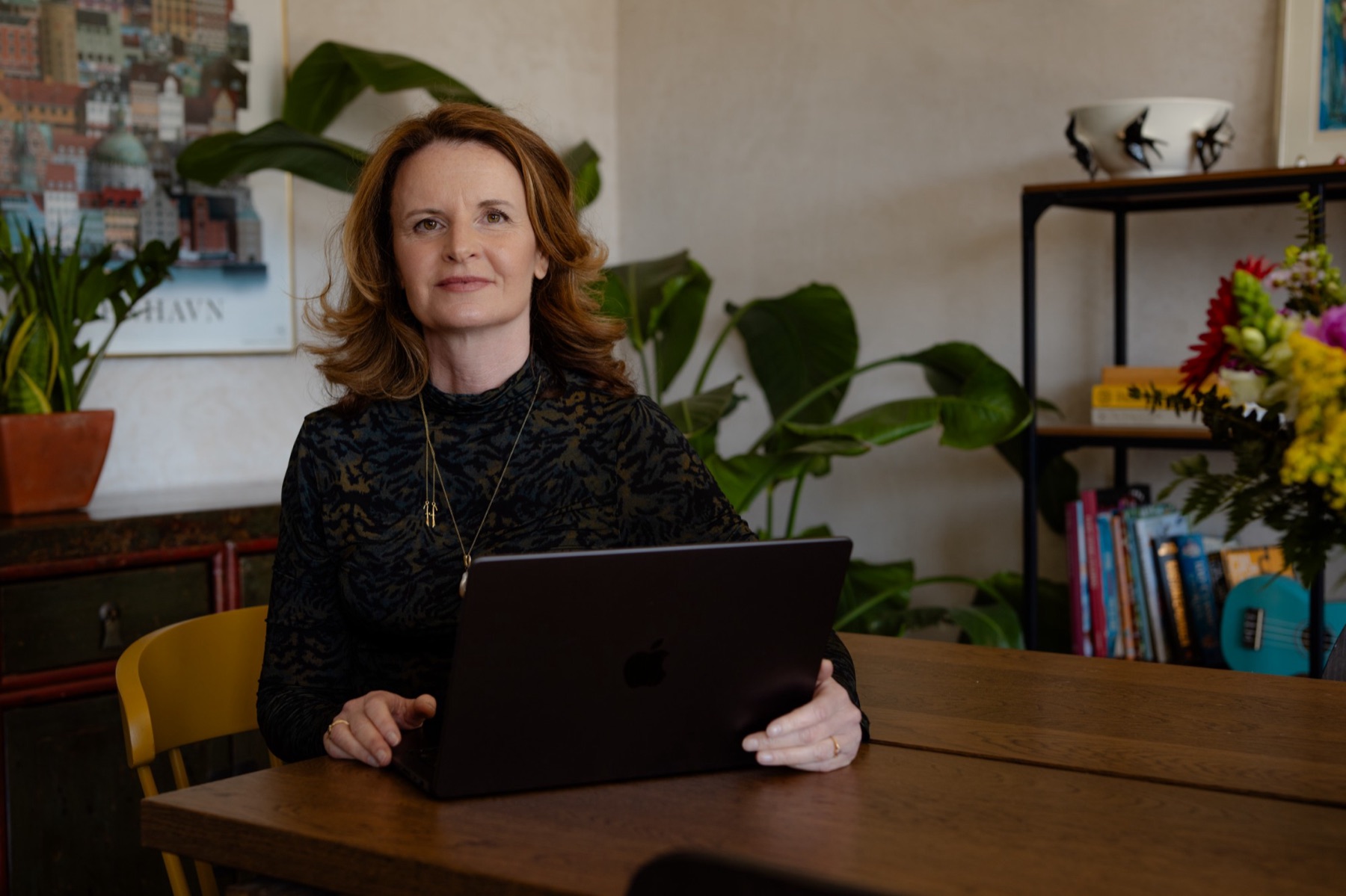 Beile Grünbaum at a wooden table with a laptop, in front of green plants and a bookshelf