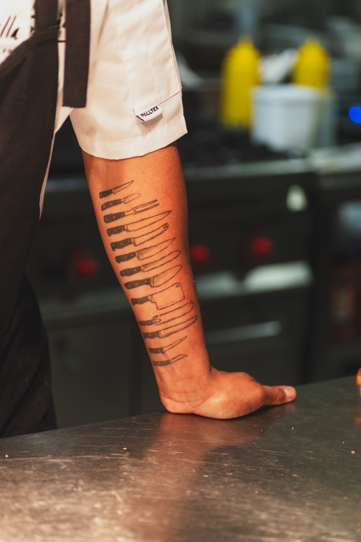 A chef’s tattooed forearm resting on a steel counter