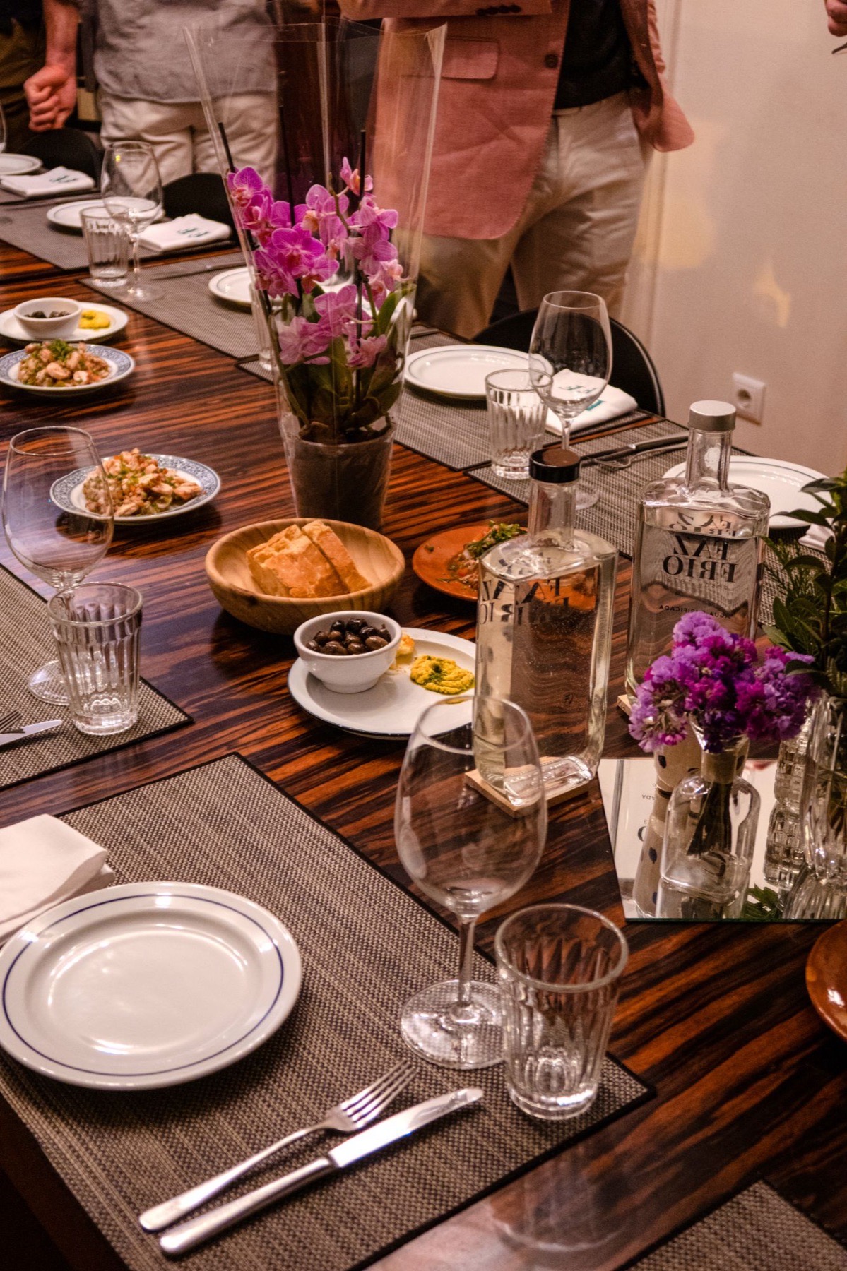 A long dinner table set with plates, glasses and flowers as guests gather