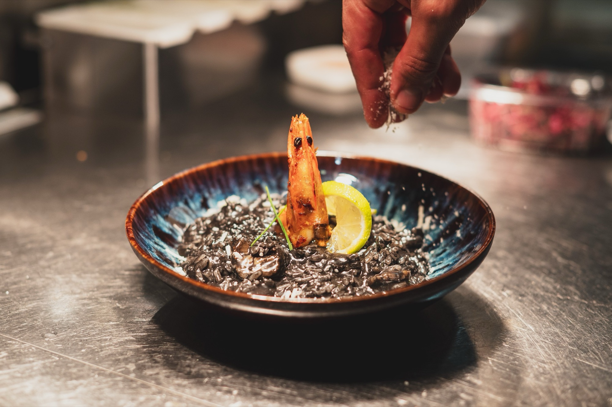 Black risotto in a blue bowl being finished by the chef's hands